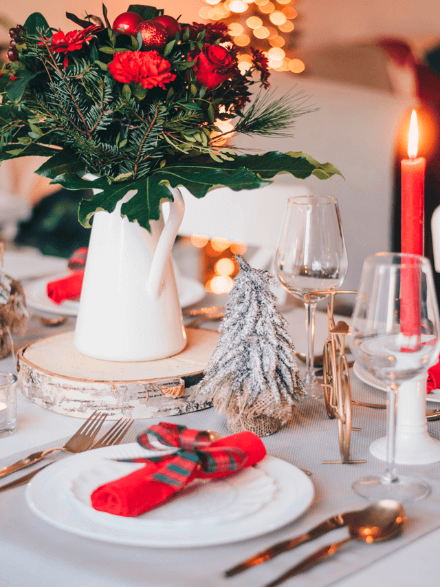 Jarra com folhagens, rosas e enfeites natalinos sobre a mesa da Ceia de Natal. Há utensílios, velas e guardanapos de tecido.